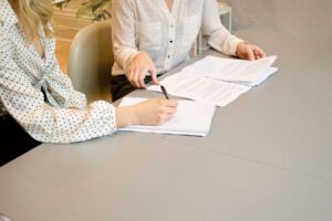 Two people sit at a table with documents spread out in front of them. One person is pointing at a document labeled "fape waiver" with a pen while the other takes notes meticulously.