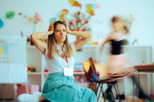A woman sitting on the floor of a colorful classroom with her hands on her head, struggling with classroom management, while a child runs around in the background displaying disruptive behavior.