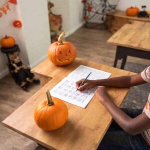 A person writes in a notebook at a wooden desk with two pumpkins, including halloween worksheets adorned with festive designs. A carved jack-o'-lantern grins nearby, while halloween decorations add a spooky charm to the scene.