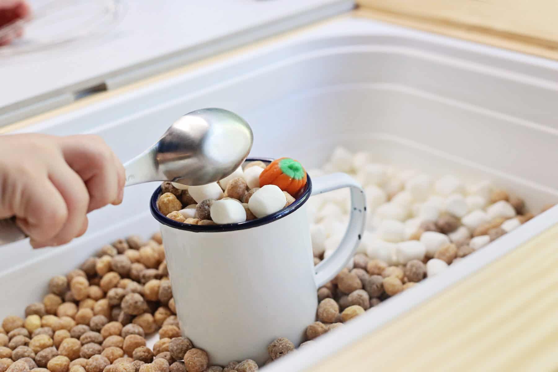 A hand uses a scoop to fill a white mug with round cereal pieces, marshmallows, and pumpkin-shaped candy in this delightful pumpkin spice sensory bin. The mug rests in a container brimming with more cereal and marshmallows.