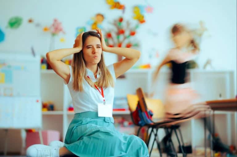 Teacher sitting at desk with head in hands looking overwhelmed while a student runs across the classroom in the background, illustrating classroom disruption and teacher stress.