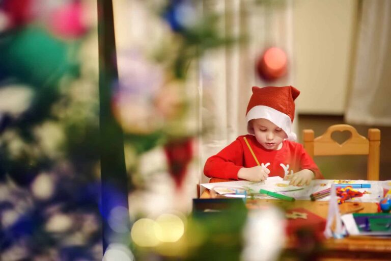 A child in a santa hat colors christmas coloring pages at a table, surrounded by art supplies, with a blurred christmas tree in the foreground.