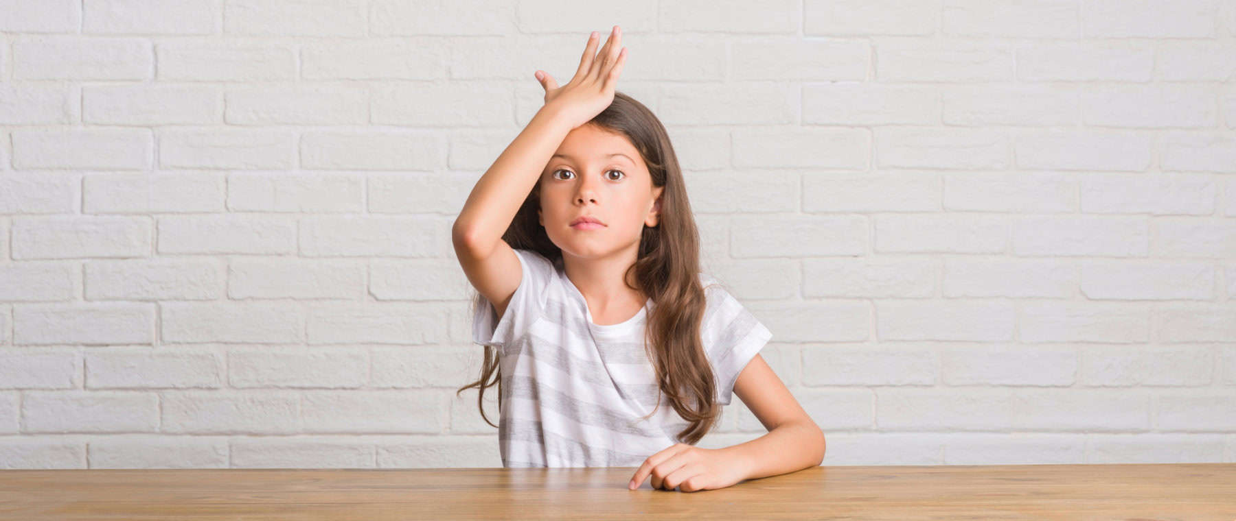 40 working memory accommodations for a 504 or iep 1 A young girl with long hair sits at a table against a white brick wall, resting her hand on her forehead, as if pondering how accommodations might aid her working memory.
