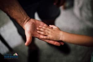 An adult hand and a child's hand touch palm to palm against a neutral background, symbolizing connection in education. The lower left corner features the logo "a day in our shoes.
