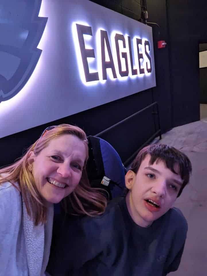 Two people smile near a large illuminated "eagles" sign in an indoor setting, as they introduce a disabled child to the vibrant atmosphere, ensuring everyone feels included in the joyful moment.