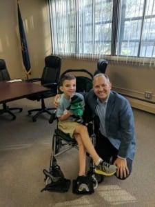 A man kneels, smiling warmly next to his disabled son who holds a stuffed toy, in an office setting adorned with a flag and conference table.