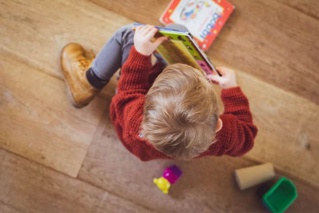 Developmental delay on an iep: what parents need to know before it’s too late 1 A child sits on the wooden floor, clad in a red sweater and brown boots, focused intently on an open book. Nearby, a collection of toys and another book lie scattered—a perfect setting to encourage exploration and potentially aid in addressing developmental delays.
