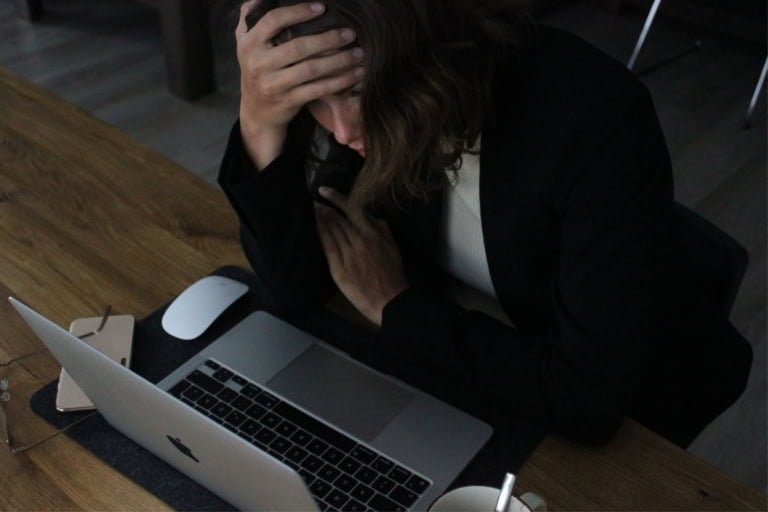 A person sits at a wooden desk, weary from the ongoing special education battle, holding their head in one hand as they look at a laptop. Nearby lie a smartphone, glasses, and a cup, silent witnesses to the struggle unfolding.