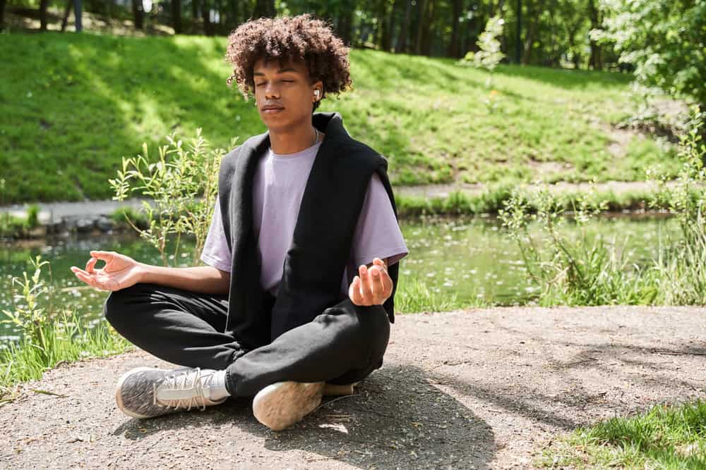 Sitting cross-legged by a tranquil pond, a person with curly hair embraces the peaceful moment. Dressed in a black hoodie and light purple shirt, they meditate with eyes closed and earbuds in, exploring whether mindfulness really works in their quest for inner peace.