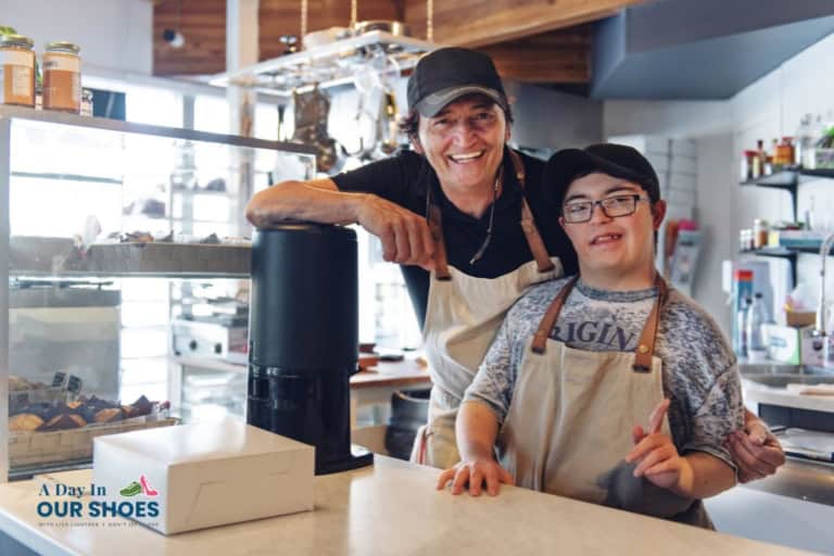 Two people wearing aprons and hats stand behind a bakery counter, smiling at the camera. One is making a peace sign. Pastries and a white box are visible on the counter, reflecting the community spirit supported by the administration for community living (acl).