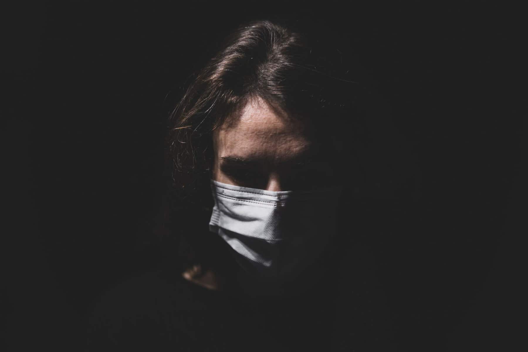 A person wearing a face mask is partially illuminated against a dark background.
