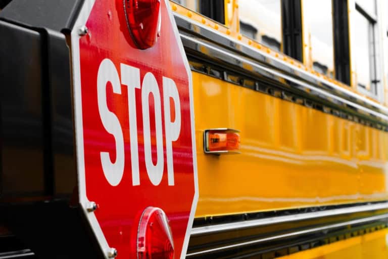 Close-up of a red octagonal stop sign mounted on the side of a yellow school bus, with visible side mirror and panel details—a small but vital measure to make schools better and keep students safe.