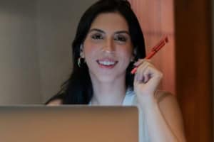 A person is sitting indoors, smiling as they hold a red pen and review a laptop screen, perhaps preparing a prior written notice.