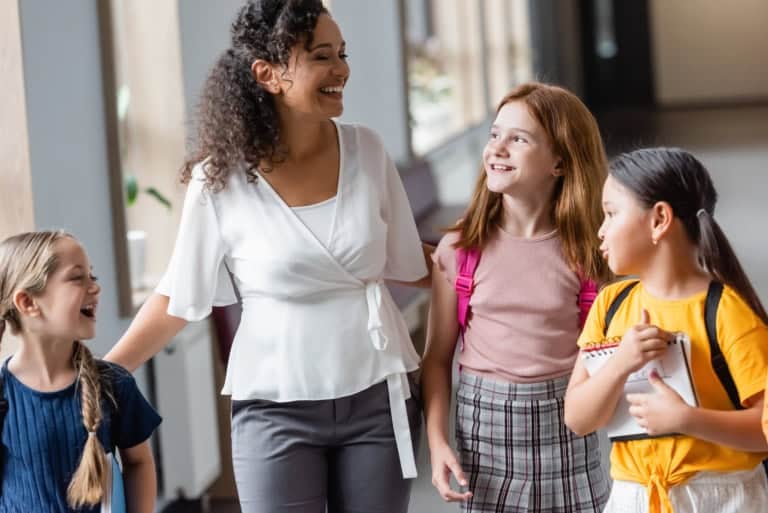 A teacher and three young students walk together in a hallway, discussing strategies for learning. The teacher is smiling as the students carry books and wear backpacks, eager to build their self-advocacy skills.