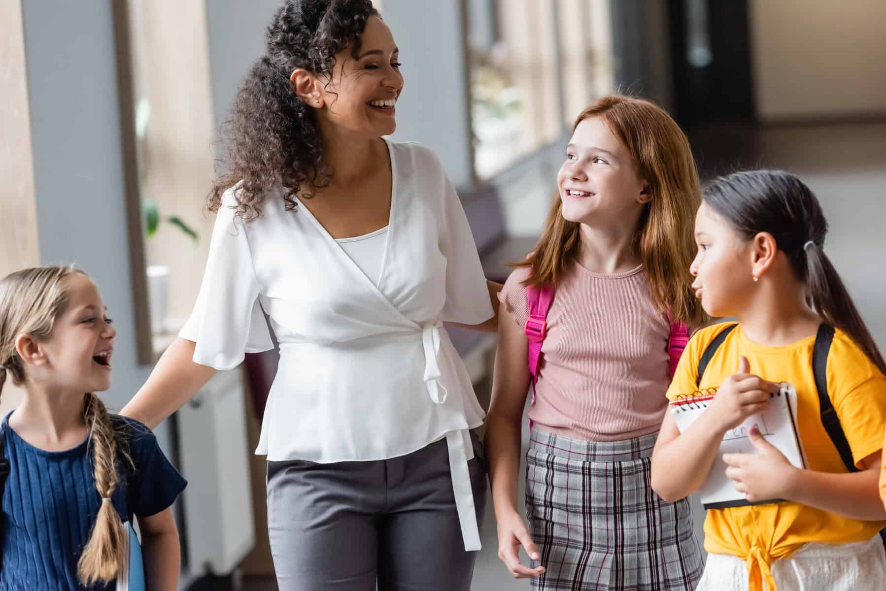 A teacher and three young students walk together in a hallway, discussing strategies for learning. The teacher is smiling as the students carry books and wear backpacks, eager to build their self-advocacy skills.