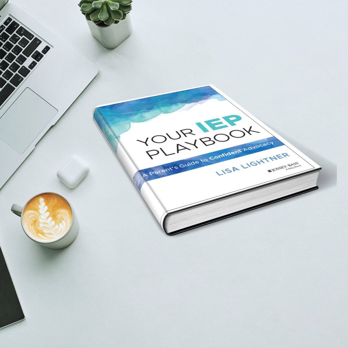 A book titled "Your IEP Playbook" by Lisa Lightner on a white desk with a laptop, cappuccino, AirPods, and a small plant.