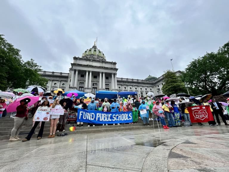 A crowd holding signs and umbrellas gathers on the steps of a government building under cloudy skies, with a prominent blue banner reading “fund public schools,” demonstrating state level advocacy tips in action.