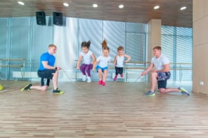 Two adults hold the ends of a jump rope while four children, participating in adaptive phys ed, jump together in a gym with wooden floors and mirrored walls, supporting iep goals for physical activity and coordination.