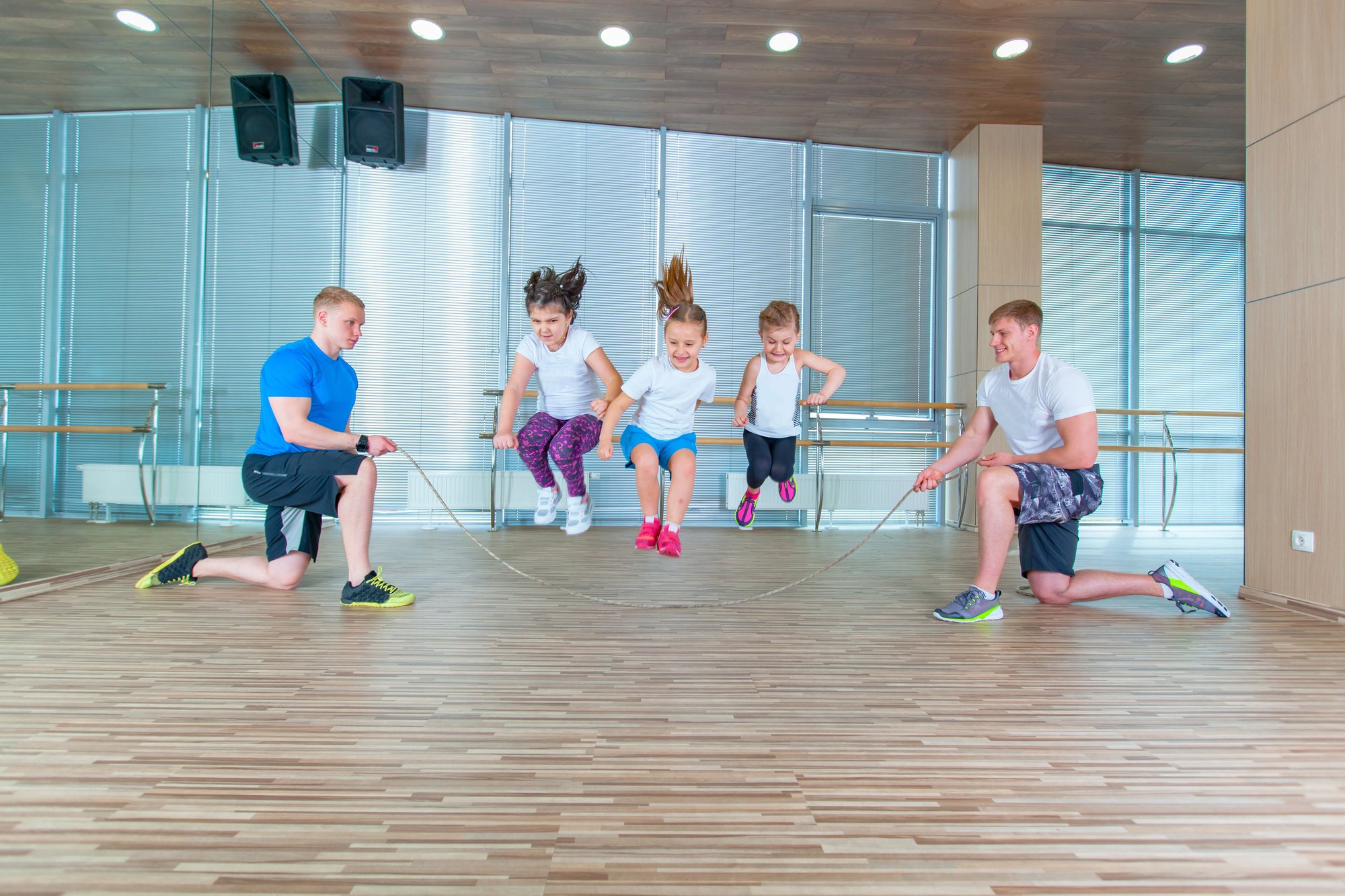 Two adults hold the ends of a jump rope while four children, participating in adaptive phys ed, jump together in a gym with wooden floors and mirrored walls, supporting iep goals for physical activity and coordination.