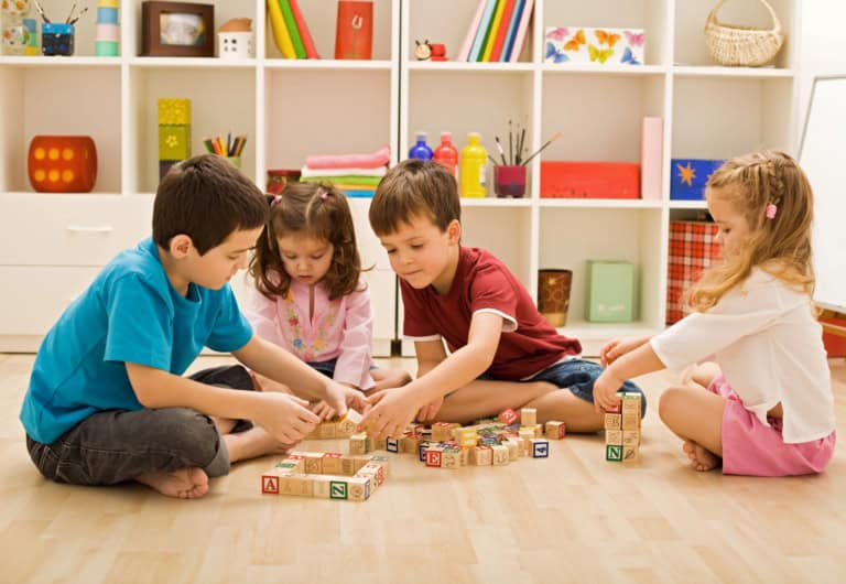 Four young children sit on the floor in a playroom, building with wooden alphabet blocks and learning social cues. Shelves with books and toys are visible in the background.