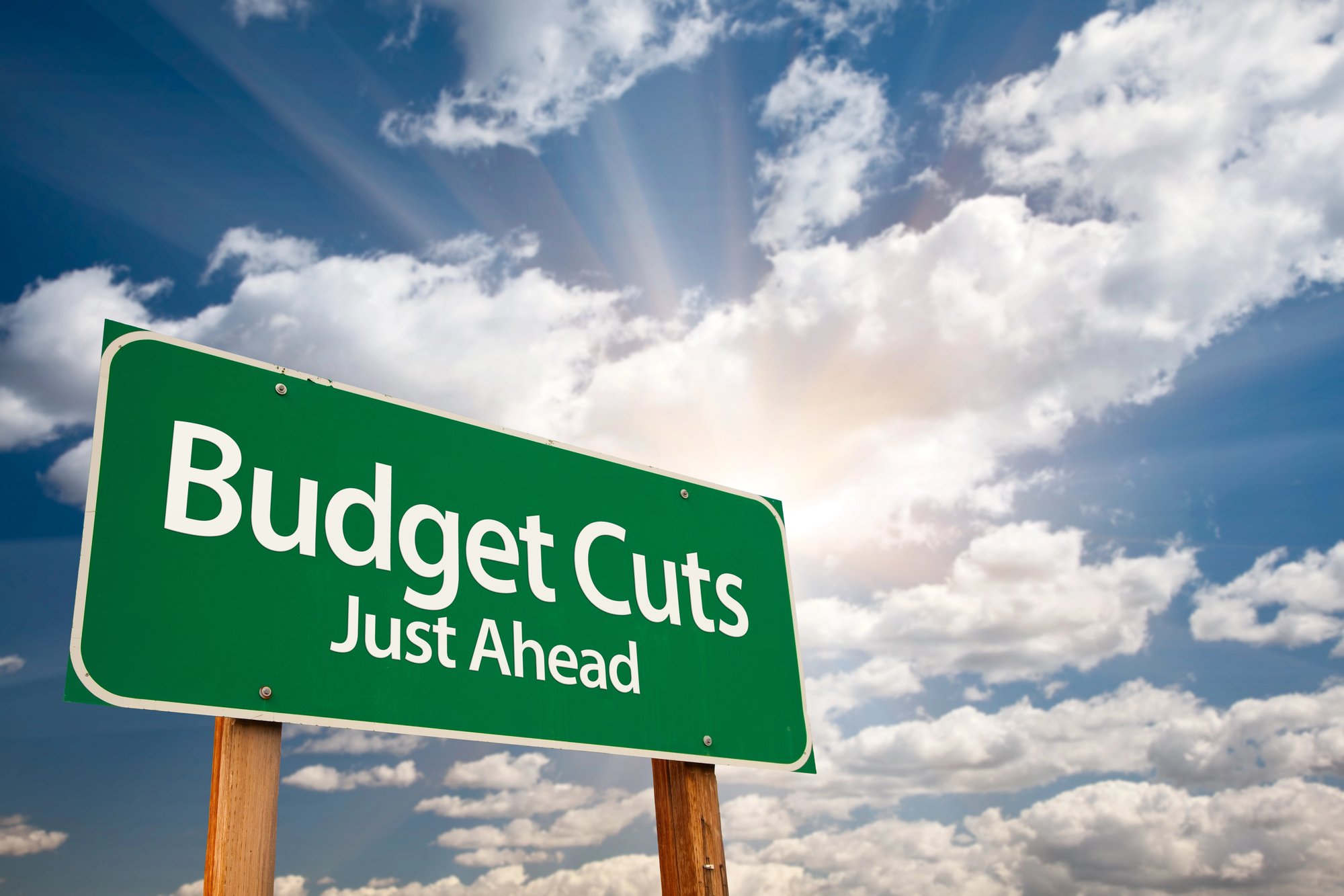 A green road sign reads “budget cuts just ahead” against a partly cloudy sky, hinting at the trump fy2026 education budget with sun rays shining through.