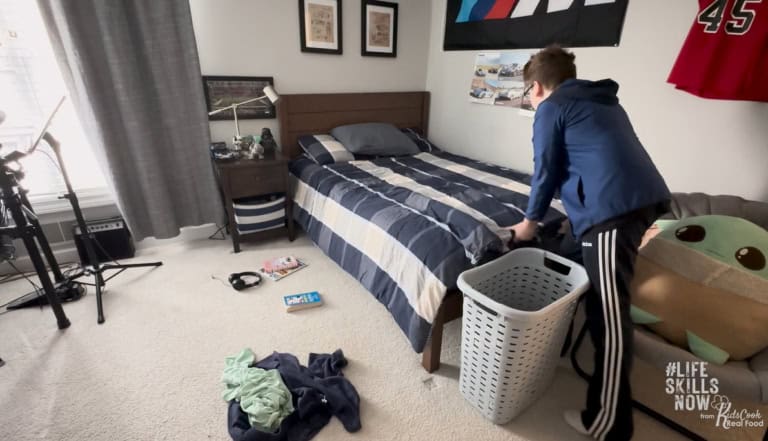 Teenage boy stands by a laundry basket in a tidy bedroom with a made bed, engaging in life skills activities as he prepares to pick up clothes from the floor.