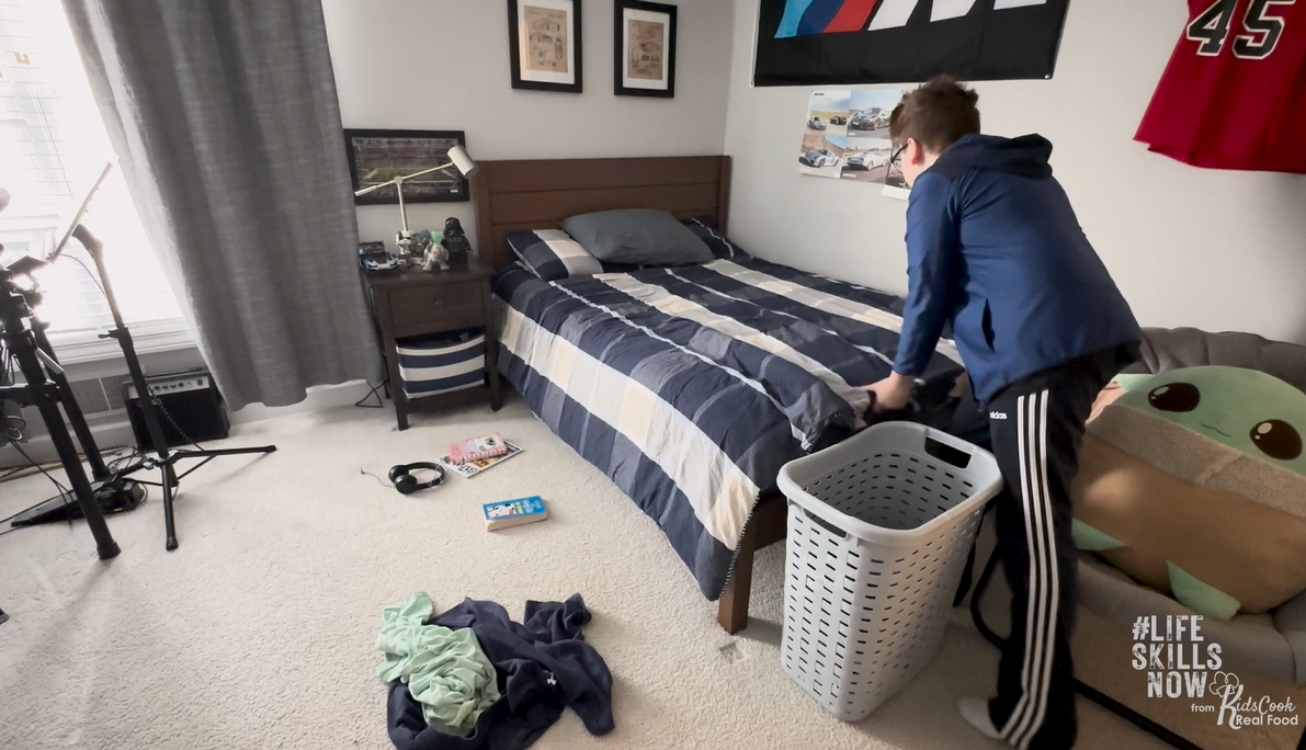 Teenage boy stands by a laundry basket in a tidy bedroom with a made bed, engaging in life skills activities as he prepares to pick up clothes from the floor.