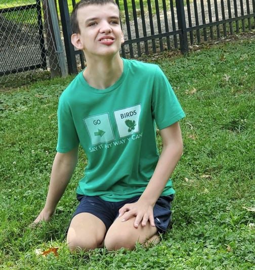 A person in a green "go birds" t-shirt and navy shorts kneels on grass near a black metal fence, looking slightly upward and smiling—showcasing the joyful spirit of autism families enjoying their game plan outdoors.