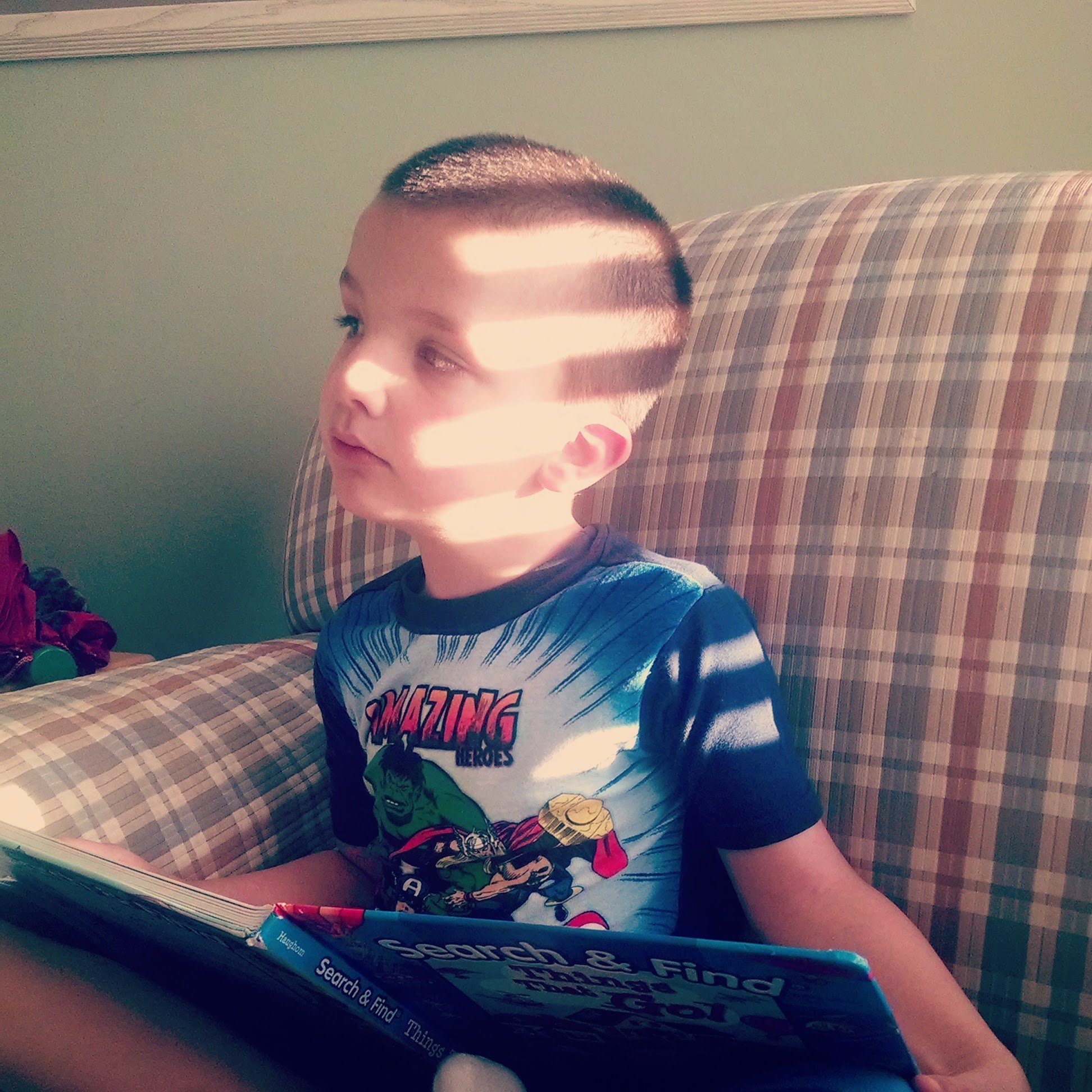 A young boy in superhero pajamas sits on a plaid armchair reading a book, sunlight streaming through blinds and casting shadows on his face—capturing the joy found when learning how to choose books for preschoolers.