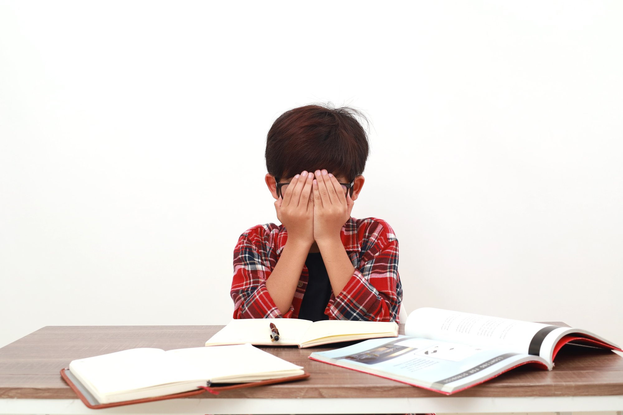 A child sitting at a desk with open books covers their face with both hands, appearing frustrated or overwhelmed, possibly in need of sensory accommodations or other classroom accommodations for sensory processing challenges.
