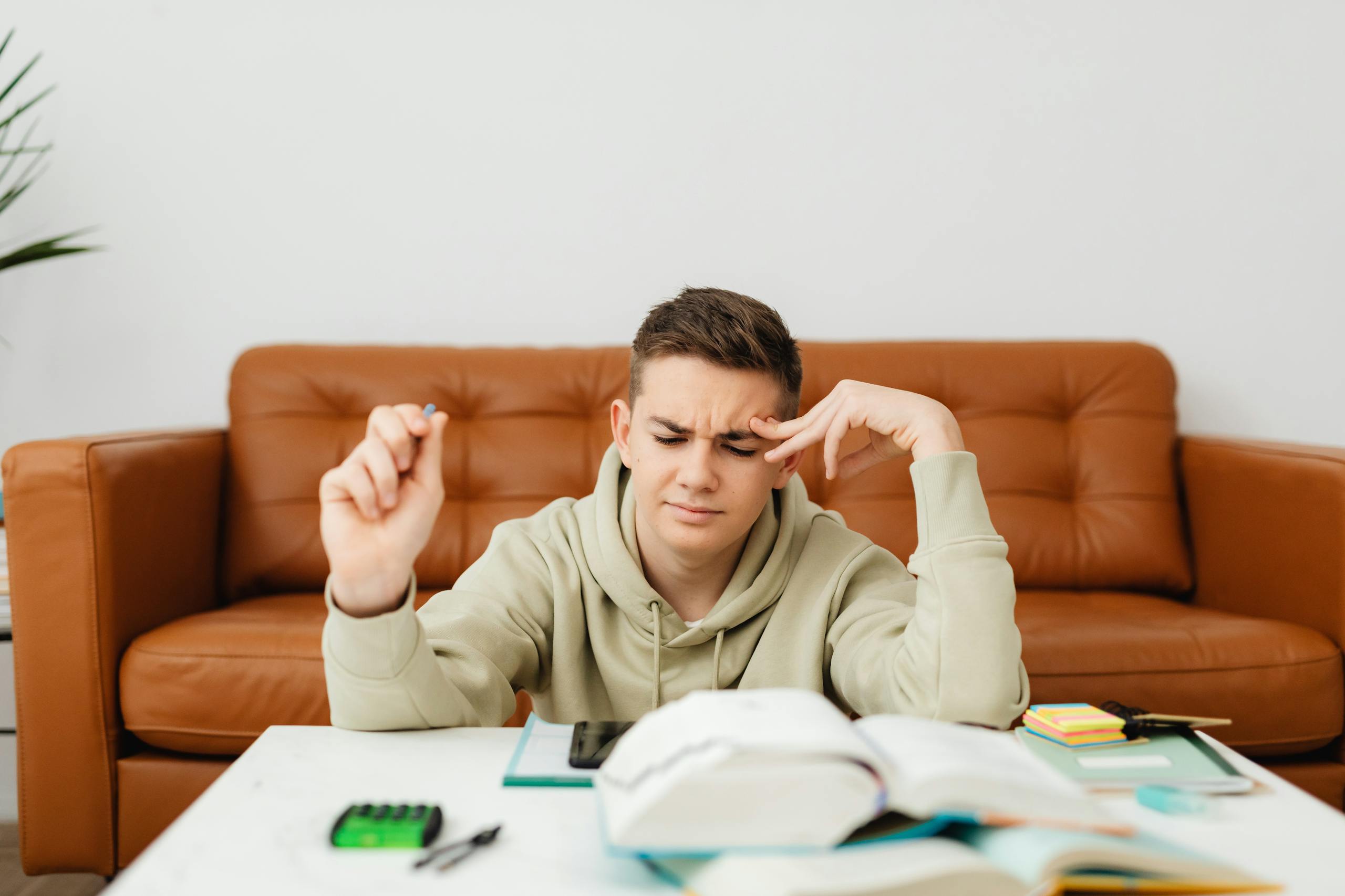 Teenage boy studying on a brown sofa, focused and thinking hard indoors.