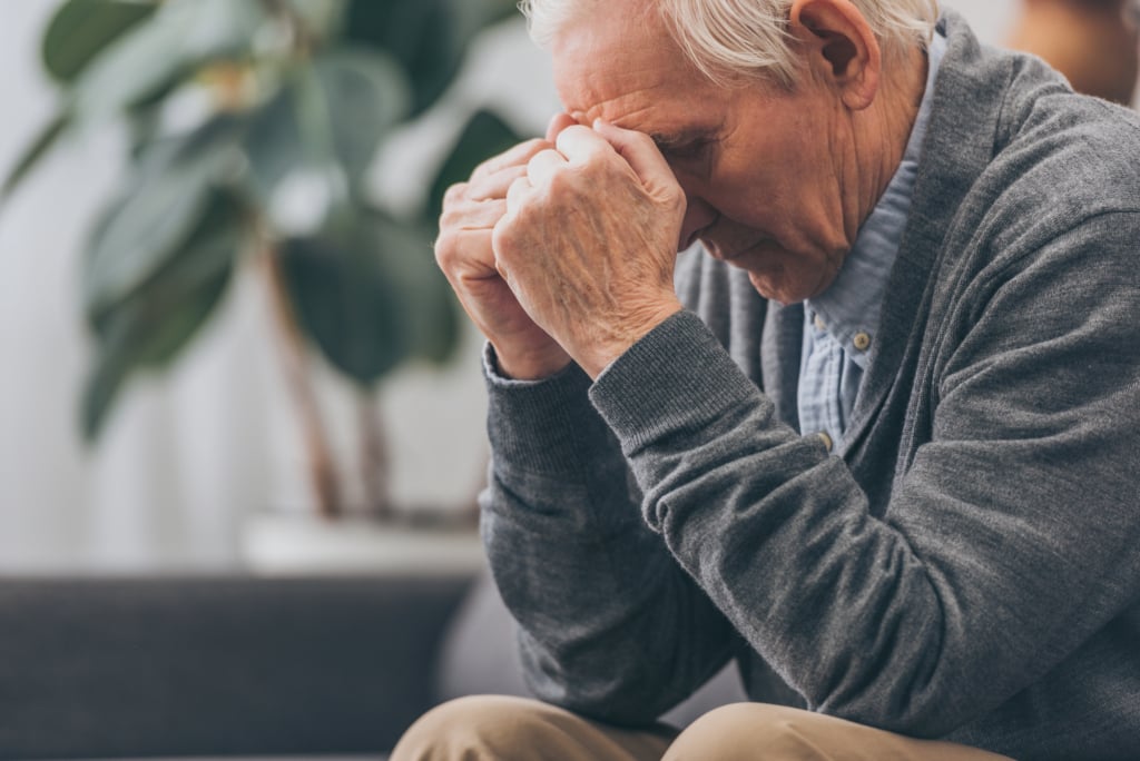 Elderly man sitting on a couch with his elbows on his knees, hands pressed to his forehead, appearing deep in thought or stressed—possibly worried about healthcare or medicare decisions.