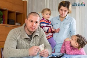 A man holds money at a table while a woman stands behind him holding a child; another child sits nearby with hands clasped, all appearing concerned about looming bills and potential bankrupt worries.