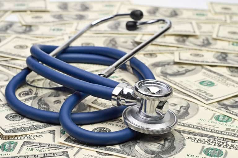 A stethoscope lies on top of scattered us hundred dollar bills, symbolizing the connection between healthcare expenses and financial costs like medicare and medicaid.
