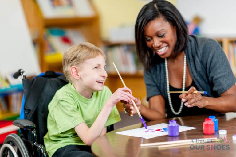 A boy in a wheelchair and a woman sit at a table, smiling and painting with brushes and colorful paints in a classroom setting, as part of his medical iep activities.
