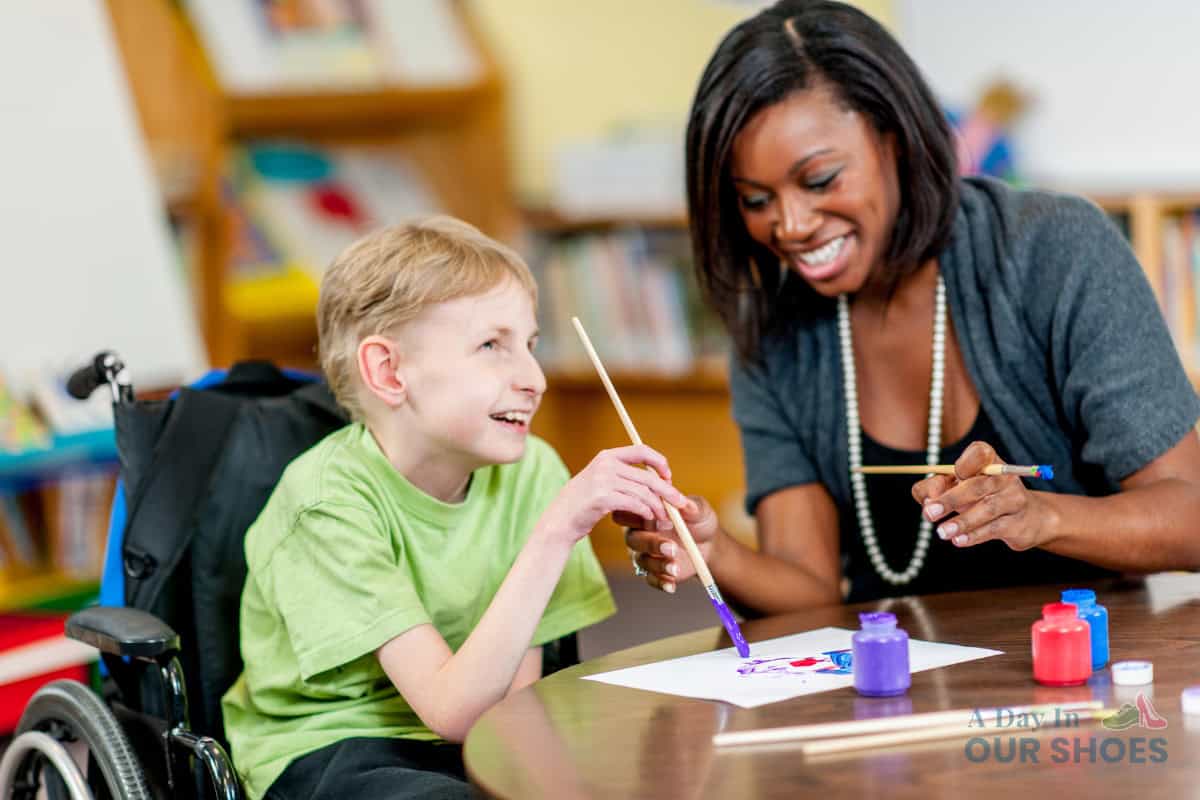 A boy in a wheelchair and a woman sit at a table, smiling and painting with brushes and colorful paints in a classroom setting, as part of his medical iep activities.
