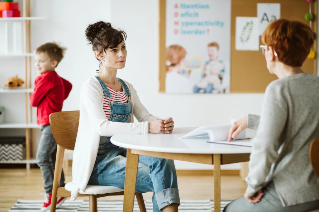 Two women sit across from each other at a table in a classroom, discussing parent observations and advocacy, while a child in a red shirt plays in the background.