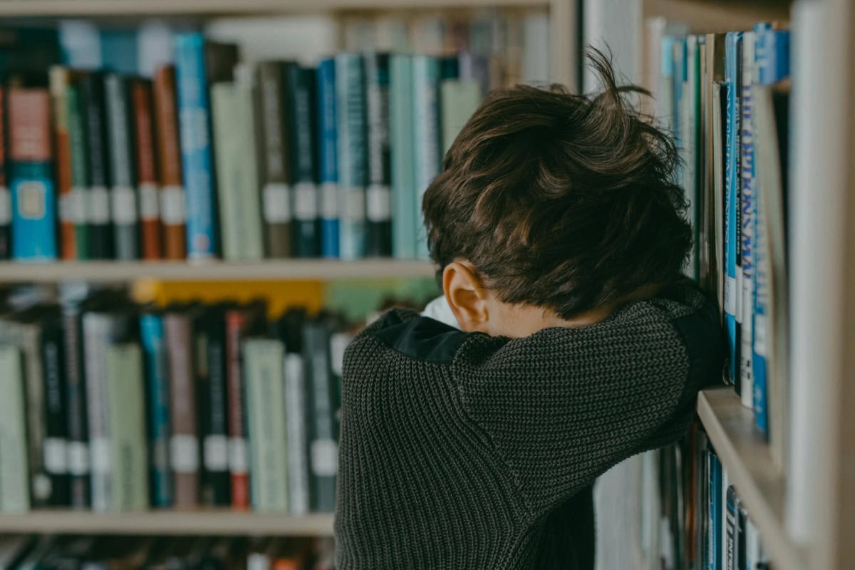 A young boy looking down, leaning against a bookshelf in a library, evoking a sense of loneliness.