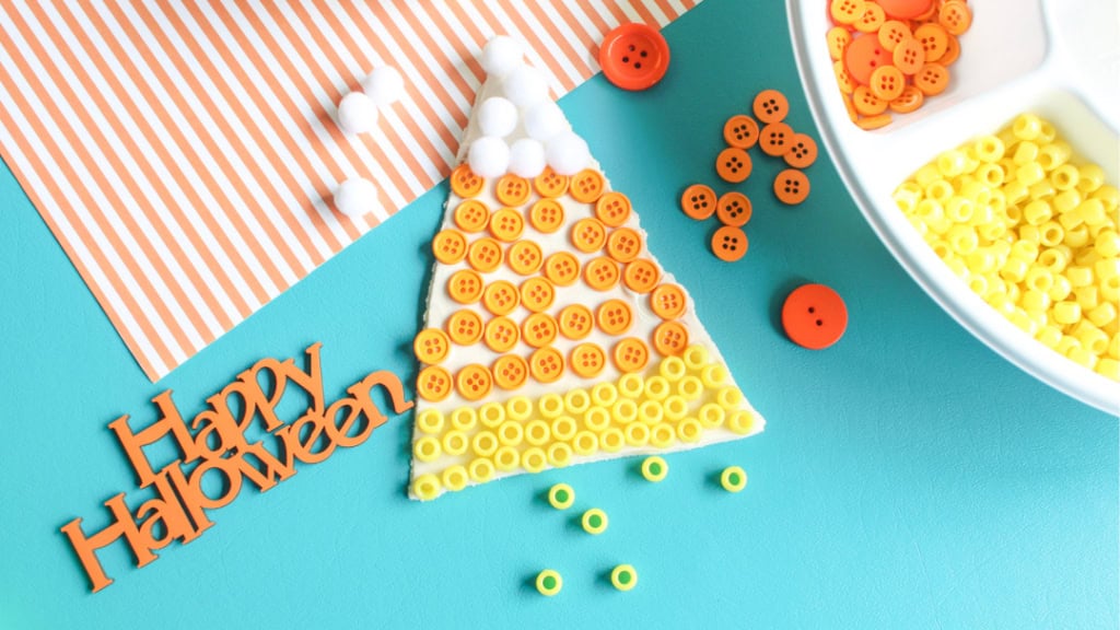A candy corn craft made with orange buttons, yellow beads, and white pom-poms sits on a blue surface next to a “happy halloween” sign and a tray of craft materials—perfect for creative halloween activities and non-food ideas.