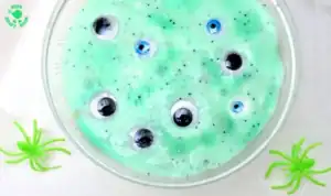 A bowl of green slime with plastic googly eyes inside, surrounded by two green toy spiders on a white surface—perfect for halloween sensory play or other non-food ideas during spooky season.