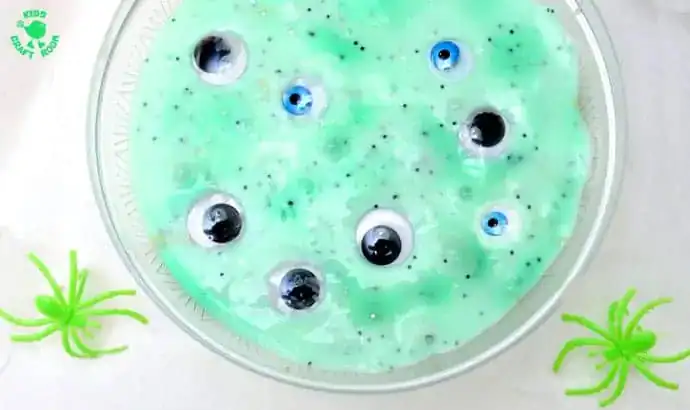 A bowl of green slime with plastic googly eyes inside, surrounded by two green toy spiders on a white surface—perfect for halloween sensory play or other non-food ideas during spooky season.