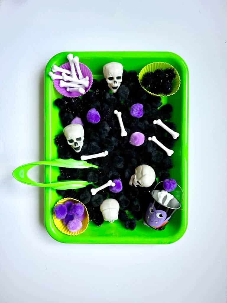 A green tray holds black pom-poms, plastic skulls and bones, purple pom-poms, cupcake liners, a small halloween cup, and a green tweezer—perfect for halloween sensory play and non-food activities.
