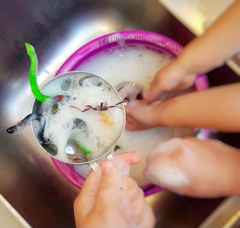 A person holds a strainer with toy snakes and foam over a sink, while two hands wash more toys in a soapy basin—an example of halloween sensory play that's perfect for non-food halloween activities.