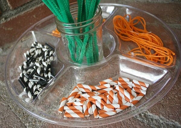 A divided tray holds black-and-white and orange-and-white paper tubes, orange yarn, and green pipe cleaners in a jar—perfect for halloween fine motor activities.