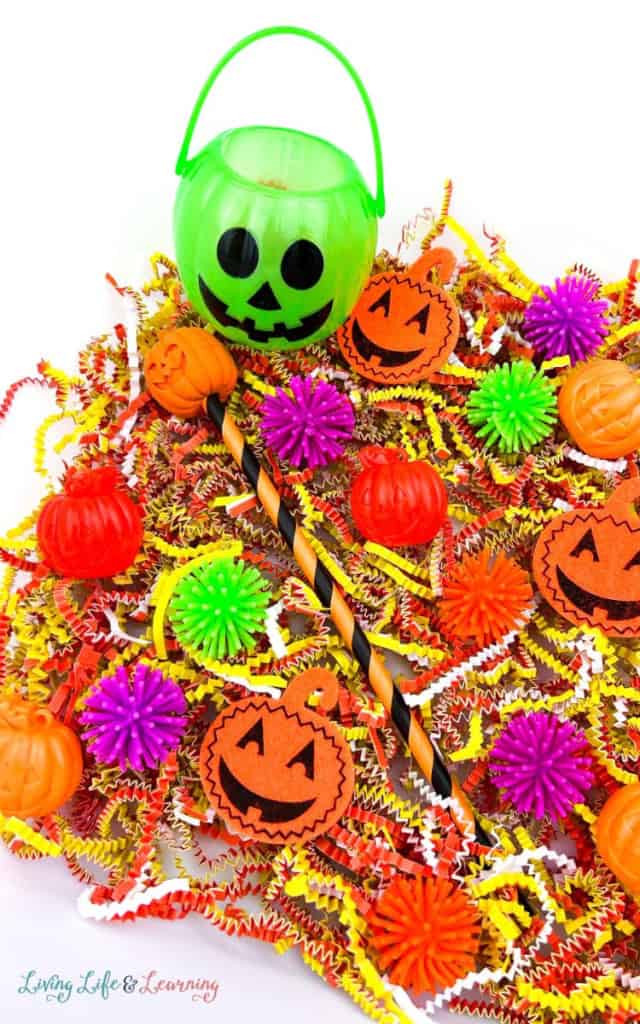A green pumpkin-shaped bucket and a halloween-themed pen are surrounded by orange, yellow, and red paper shreds, plastic pumpkins, and colorful spiky balls—perfect for non-food halloween activities or creative halloween sensory play.