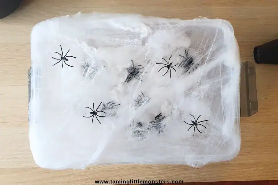 A plastic container covered in artificial spiderwebs with several fake black spiders placed on top—perfect for halloween sensory play or creative non-food ideas at your next halloween activities event.