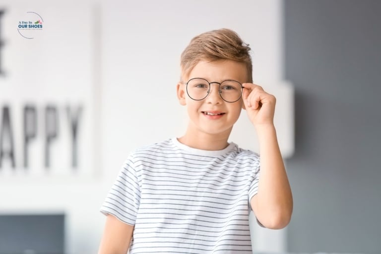 A young boy wearing adhd glasses and a white striped shirt smiles at the camera while holding his glasses with one hand indoors.