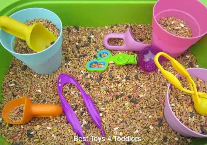 A sensory bin filled with birdseed, colorful cups, scoops, tongs, a spoon, and toy scissors—perfect for sensory play and fun kids activities.