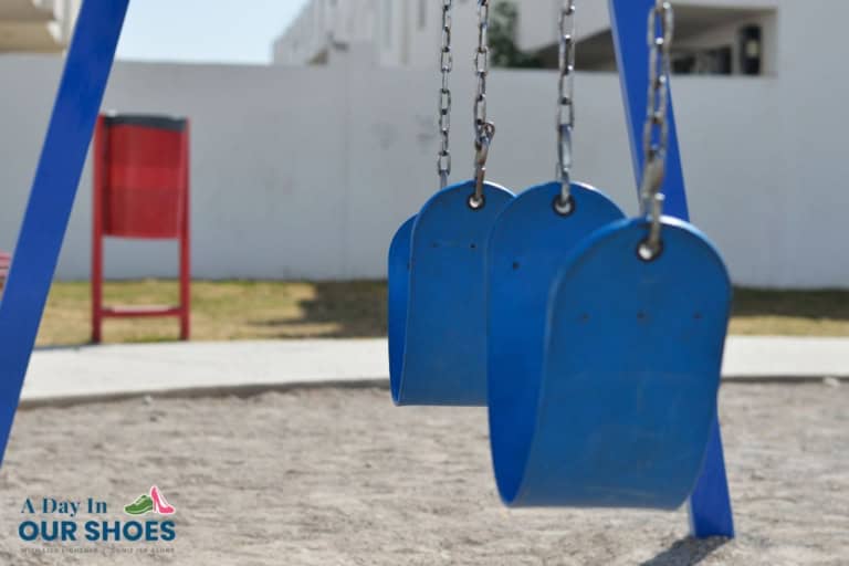 Two empty blue swings hang from chains in a playground, evoking memories of school recess punishment, with a red bench and white buildings visible in the background.