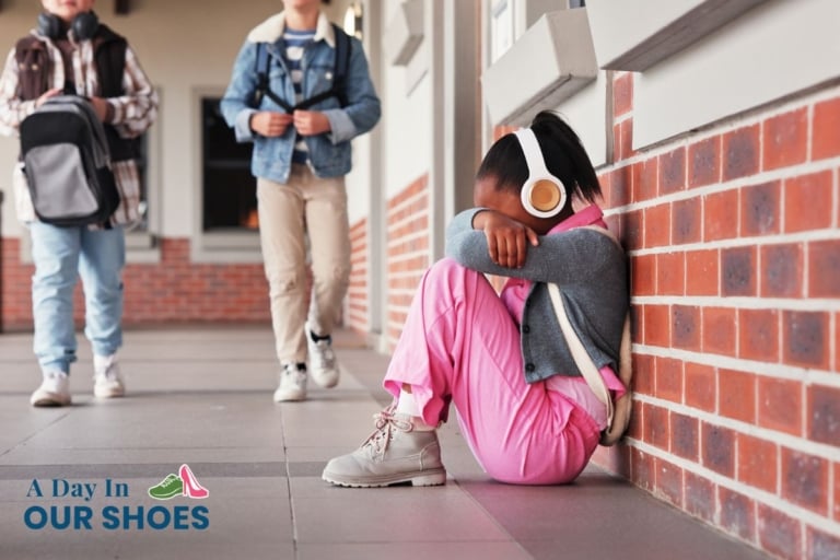 A young child wearing headphones sits on the ground against a brick wall with their head down, while other children walk by in the background.